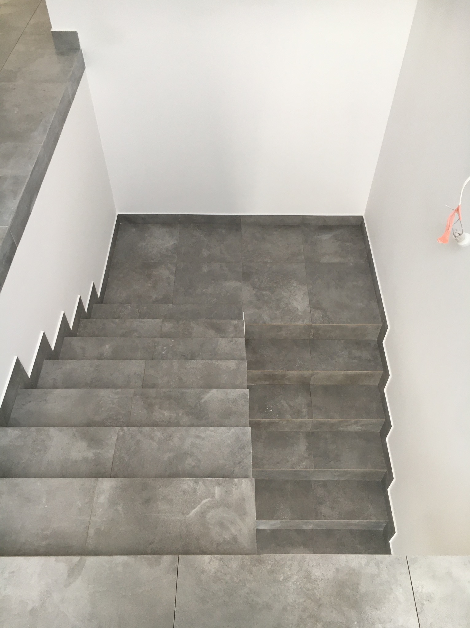 Schody wyłożone szarymi płytkami ceramicznymi, widok z góry, białe ściany, surowa żarówka z kablem w rogu.