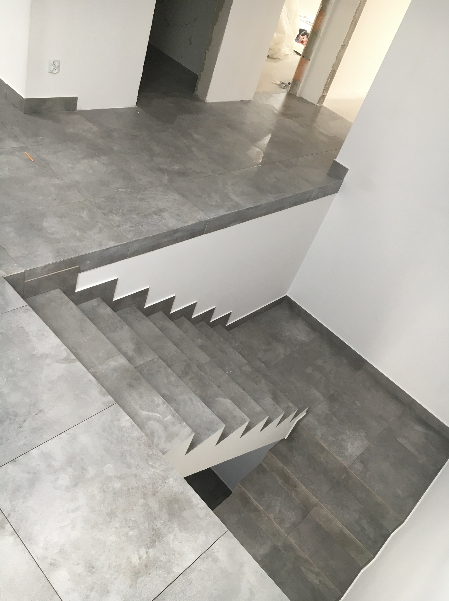 Wykończone schody z szarych płytek prowadzące do pomieszczenia z błyszczącą podłogą z płytek, widoczne gniazdko elektryczne na ścianie i fragment niezabezpieczonego otworu drzwiowego w tle.