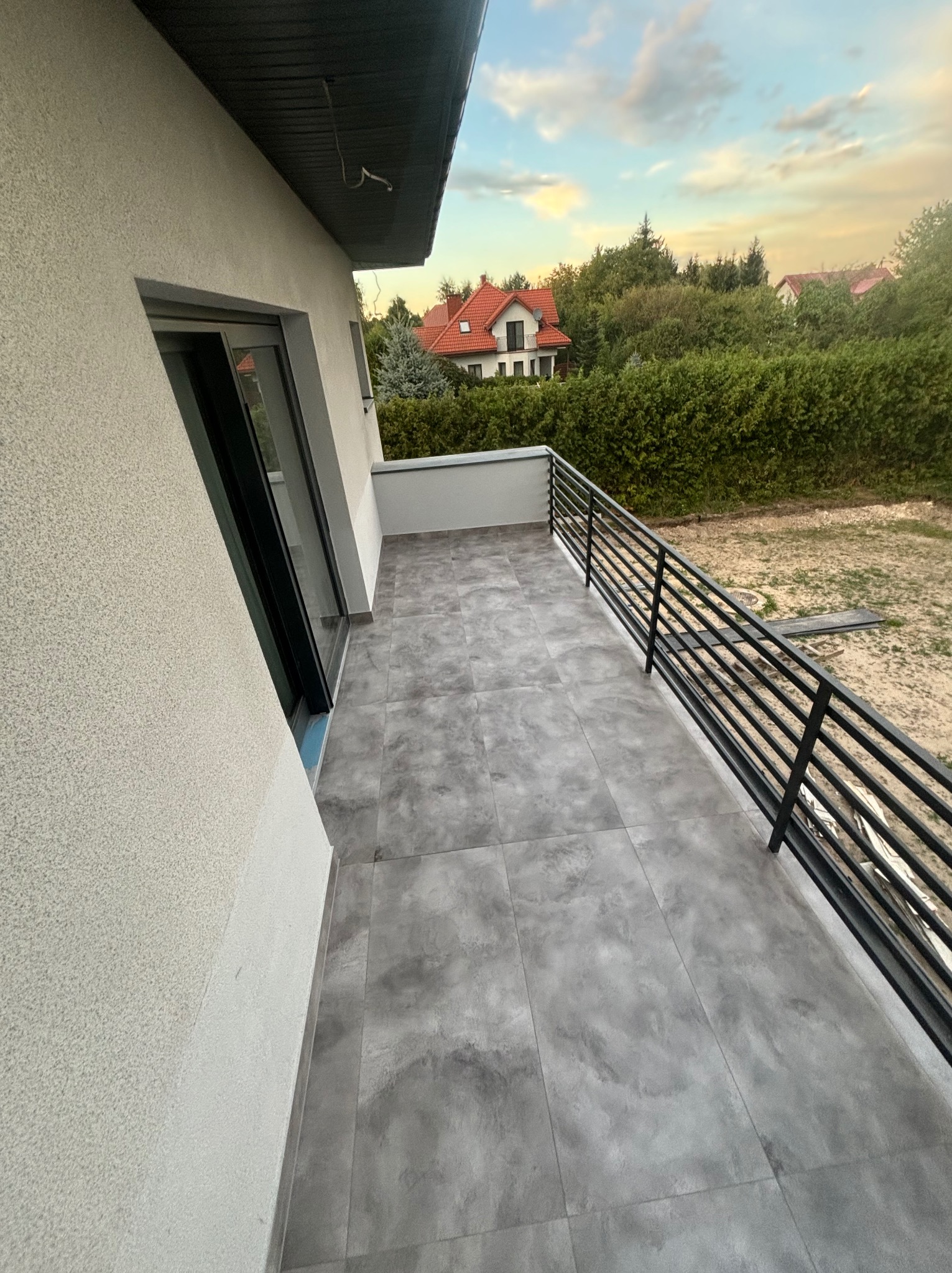 Wykończony balkon z szarą, wzorzystą terakotą i czarną, metalową balustradą na tle elewacji domu i zieleni ogrodu.