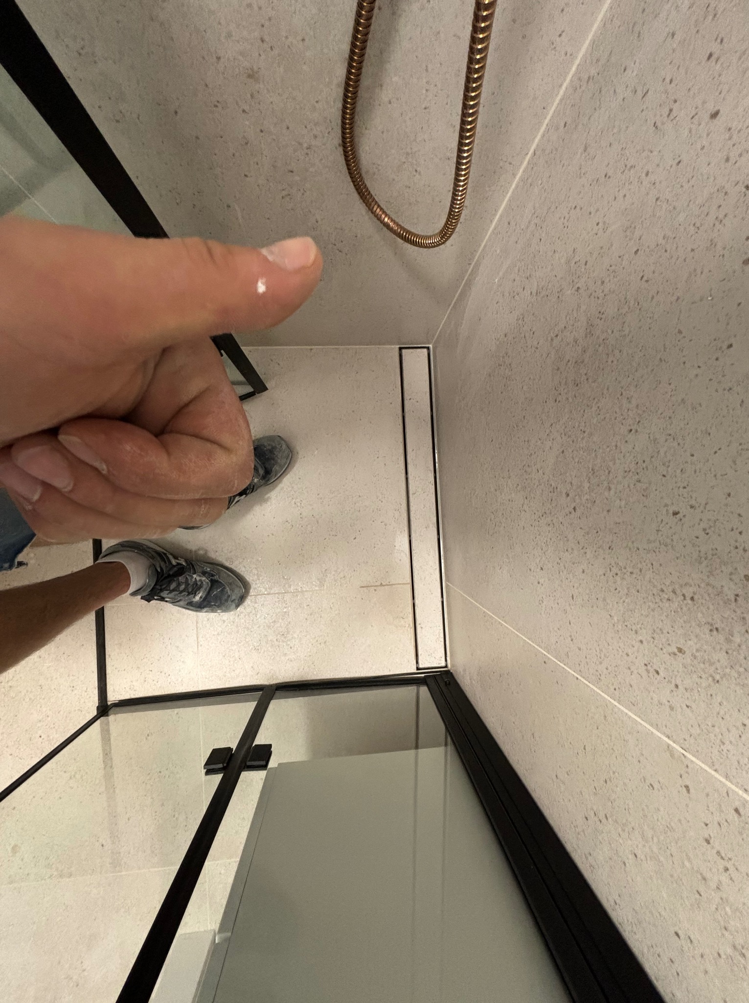 Ukończona kabina prysznicowa z odpływem liniowym, widok z góry, palec wskazujący na kroplę wody na obudowie, szare płytki na ścianach i podłodze.