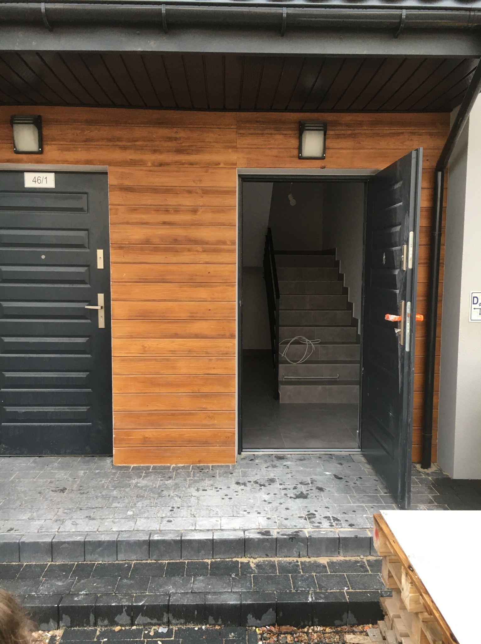 Wejście do budynku z drewnianą elewacją, czarnymi drzwiami z numerem 46/1 i otwartymi drzwiami ukazującymi schody wewnętrzne, widoczne ślady wilgoci na chodniku.