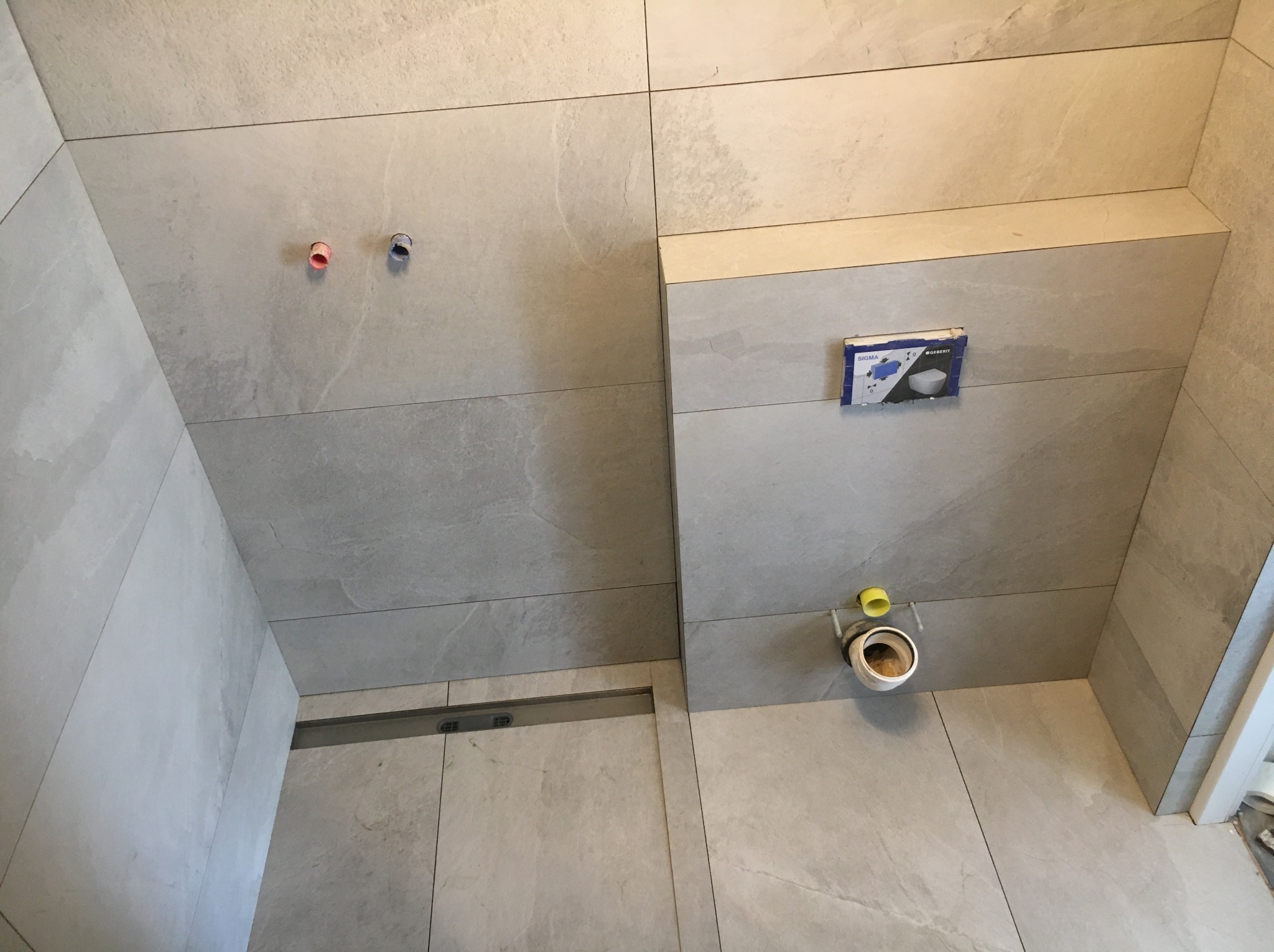 Łazienka w trakcie montażu: szare płytki wielkoformatowe, odpływ liniowy w podłodze, instalacja podtynkowa WC z widocznym kartonem producenta, wystające rury doprowadzające wodę.