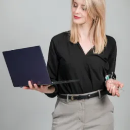 Kobieta w czarnej bluzce i szarych spodniach trzyma laptop z ciemną obudową i okulary w złotej oprawce, spoglądając w dół na laptop.