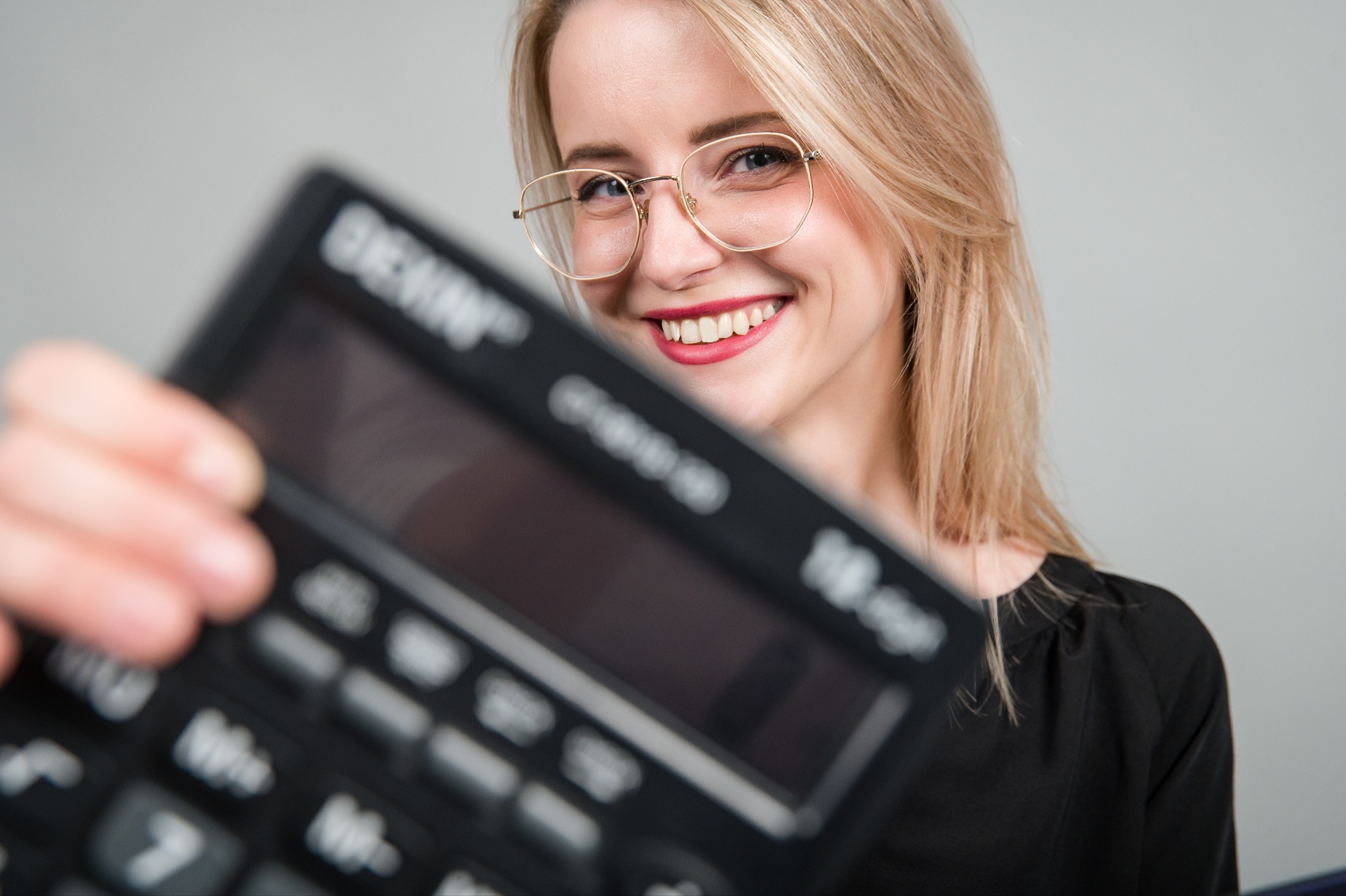 Uśmiechnięta blondynka w okularach prezentuje kalkulator, trzymając go blisko obiektywu, co powoduje efekt rozmycia tła.