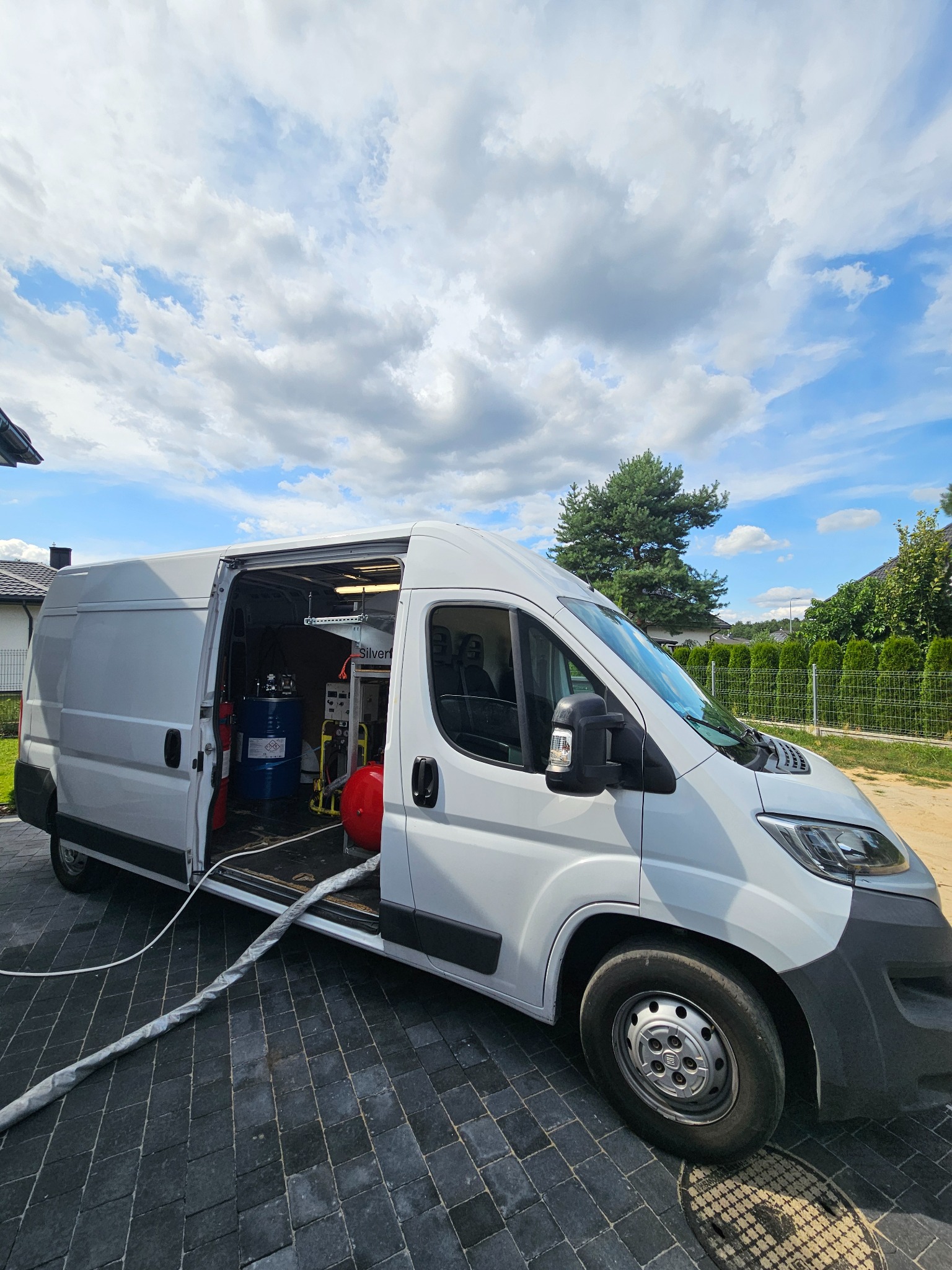Biały van z otwartymi drzwiami bocznymi, wewnątrz widoczne zbiorniki i sprzęt do natrysku, prawdopodobnie do izolacji, z pojazdu wychodzi wąż na brukowaną nawierzchnię, w tle dom i błękitne niebo...