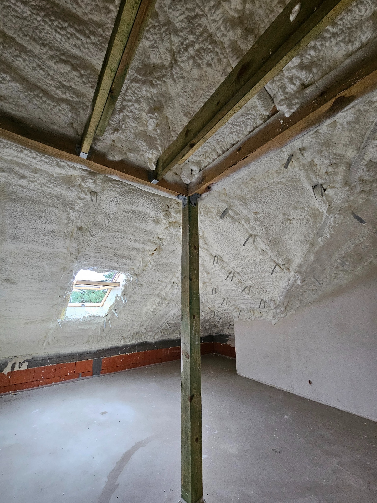 Poddasze z widoczną warstwą izolacji natryskowej z pianki, drewnianą konstrukcją więźby dachowej oraz oknem dachowym z widokiem na zieleń