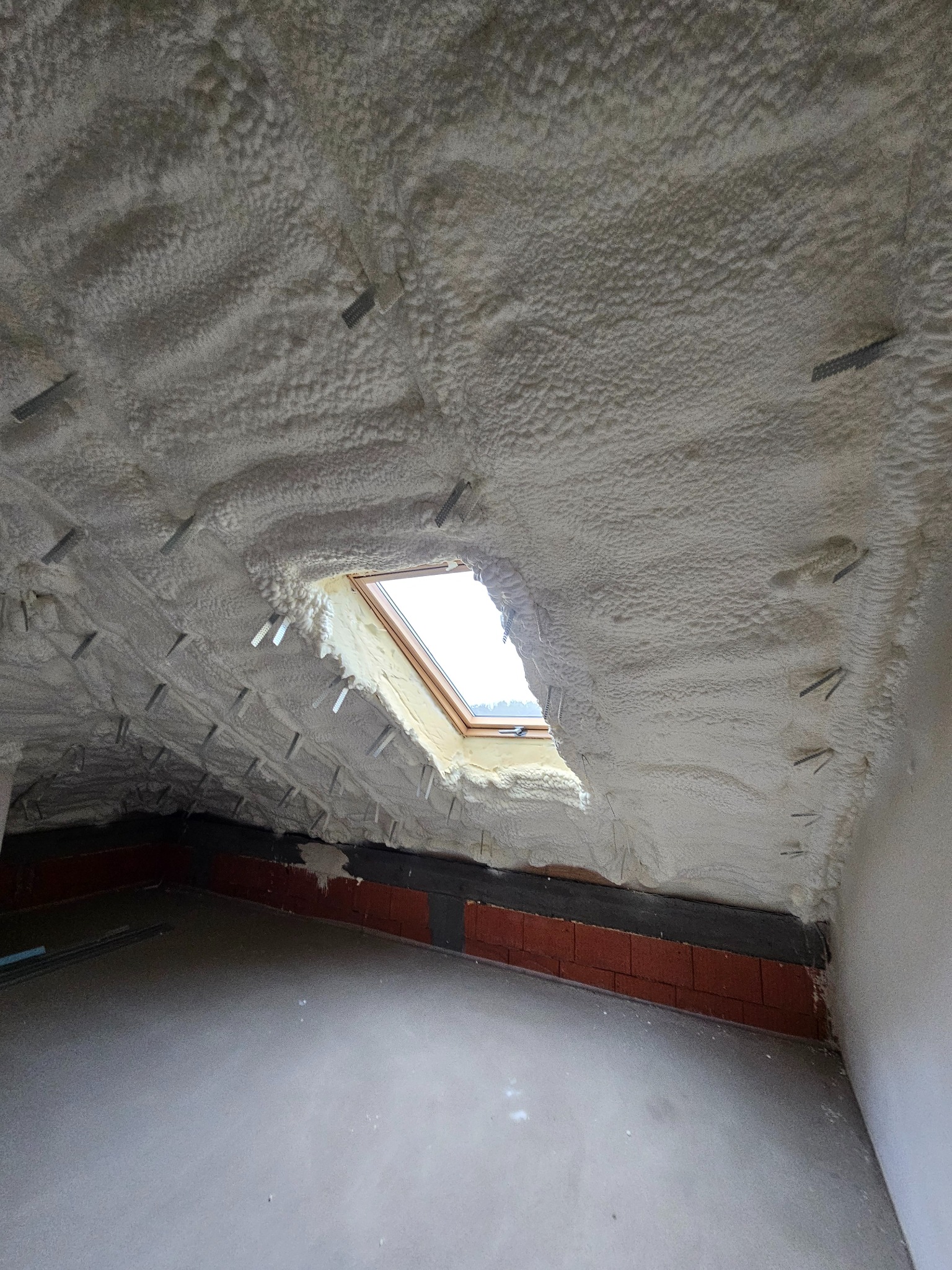 Poddasze z oknem dachowym po aplikacji izolacji pianką, widoczne metalowe elementy mocujące i fragment ściany z ceglanym wykończeniem.