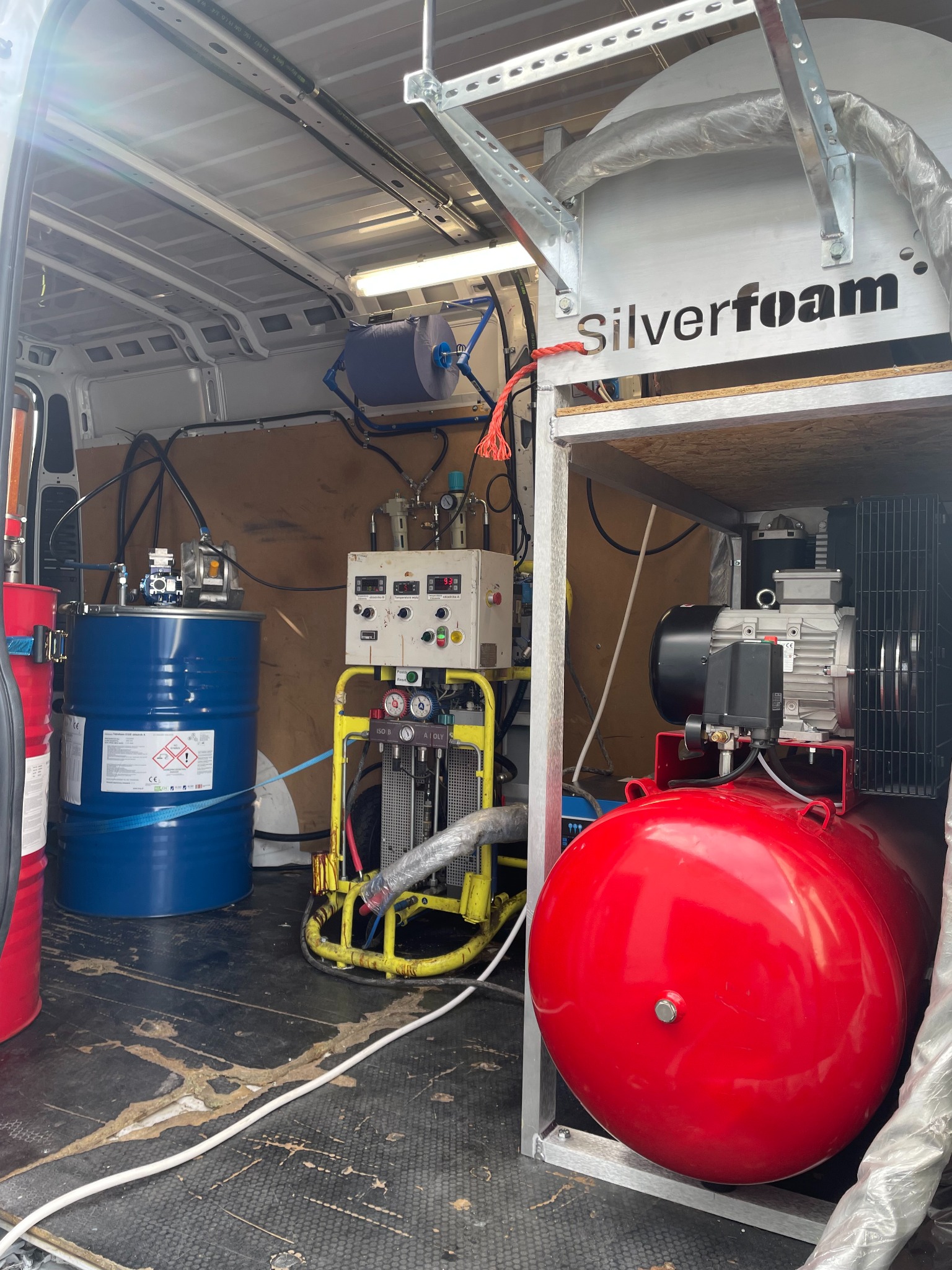 Wnętrze furgonetki z profesjonalnym sprzętem do natrysku pianki izolacyjnej: czerwony zbiornik ciśnieniowy, niebieska beczka z chemikaliami, żółta rama z manometrami i przewodami, oraz rolka...