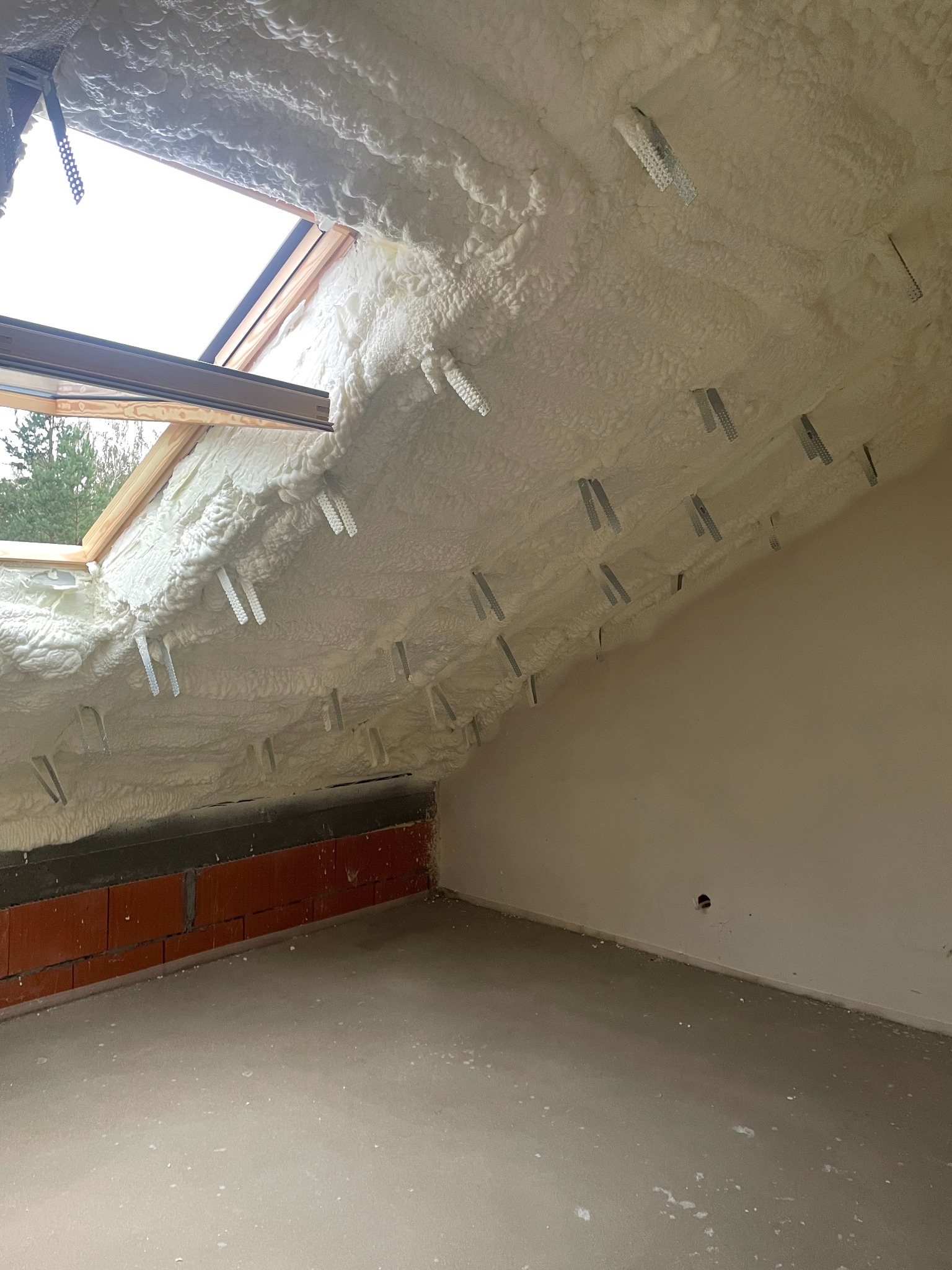 Poddasze z oknem dachowym po natrysku izolacji piankowej, widoczne metalowe uchwyty mocujące, surowy mur z cegły i betonowa posadzka.