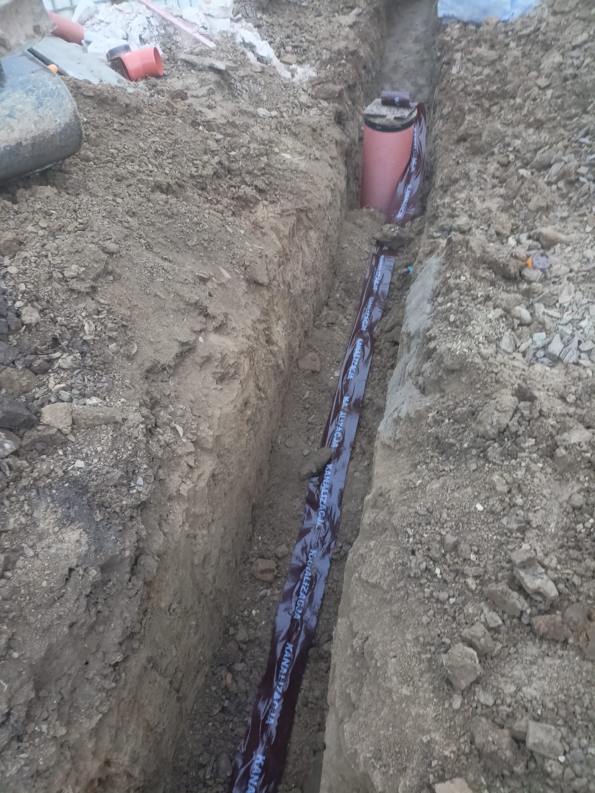 Wykop pod instalacje z rurą kanalizacyjną w brązowej folii ochronnej z napisem 'Kanalizacja' oraz studzienką rewizyjną