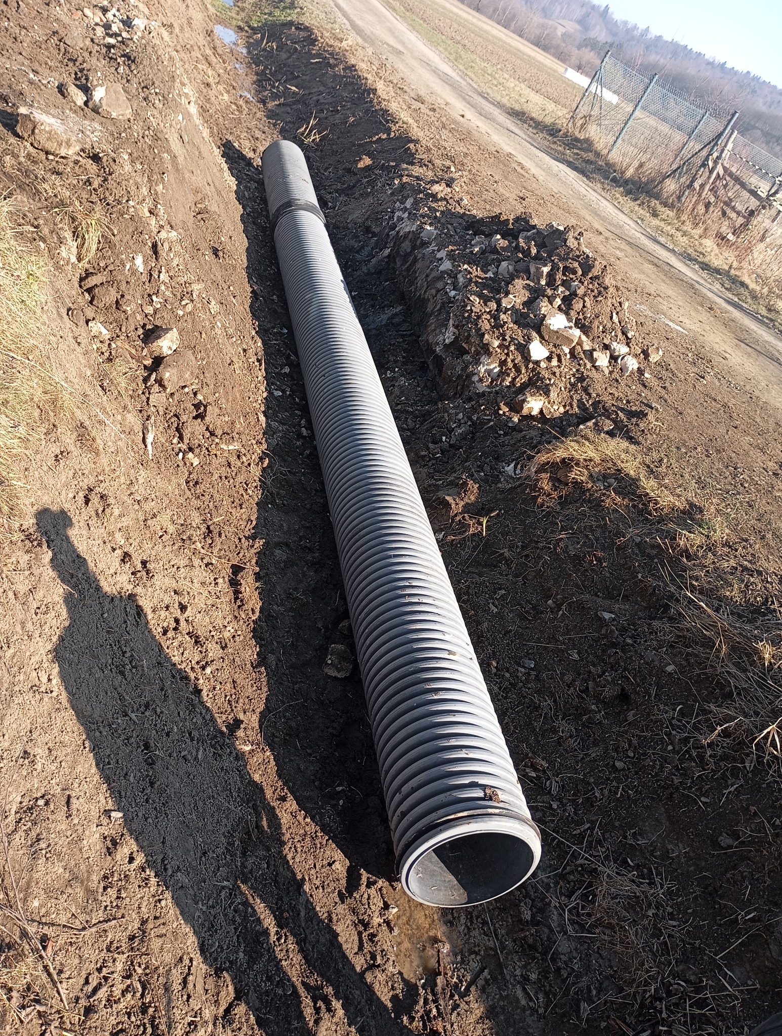 Długa, karbowana rura drenażowa leżąca w wykopie, z widocznym cieniem osoby wykonującej zdjęcie, obok usypana ziemia i w tle widok na pole i ogrodzenie.