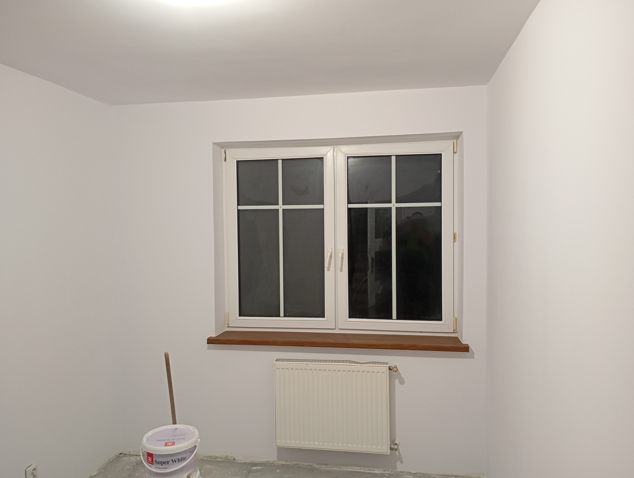 Wnętrze pokoju po malowaniu ścian na biało, widoczne okno z drewnianym parapetem, grzejnik i wiadro z farbą Super White.