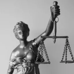 Czarno-biała statuetka Temidy, bogini sprawiedliwości, z wagą w dłoni, ukazana w dynamicznym ujęciu z dołu, podkreślającym jej majestat.