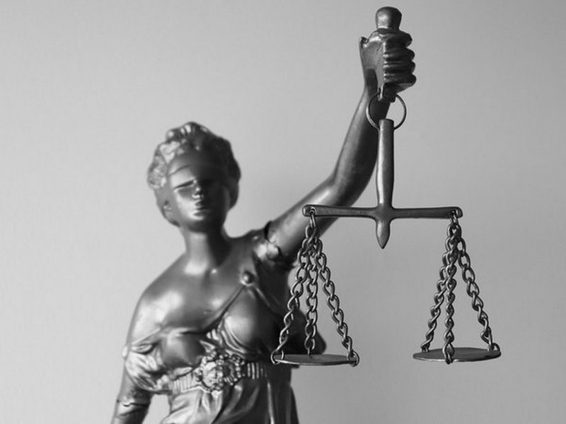 Czarno-biała statuetka Temidy, bogini sprawiedliwości, z wagą w dłoni, ukazana w dynamicznym ujęciu z dołu, podkreślającym jej majestat.