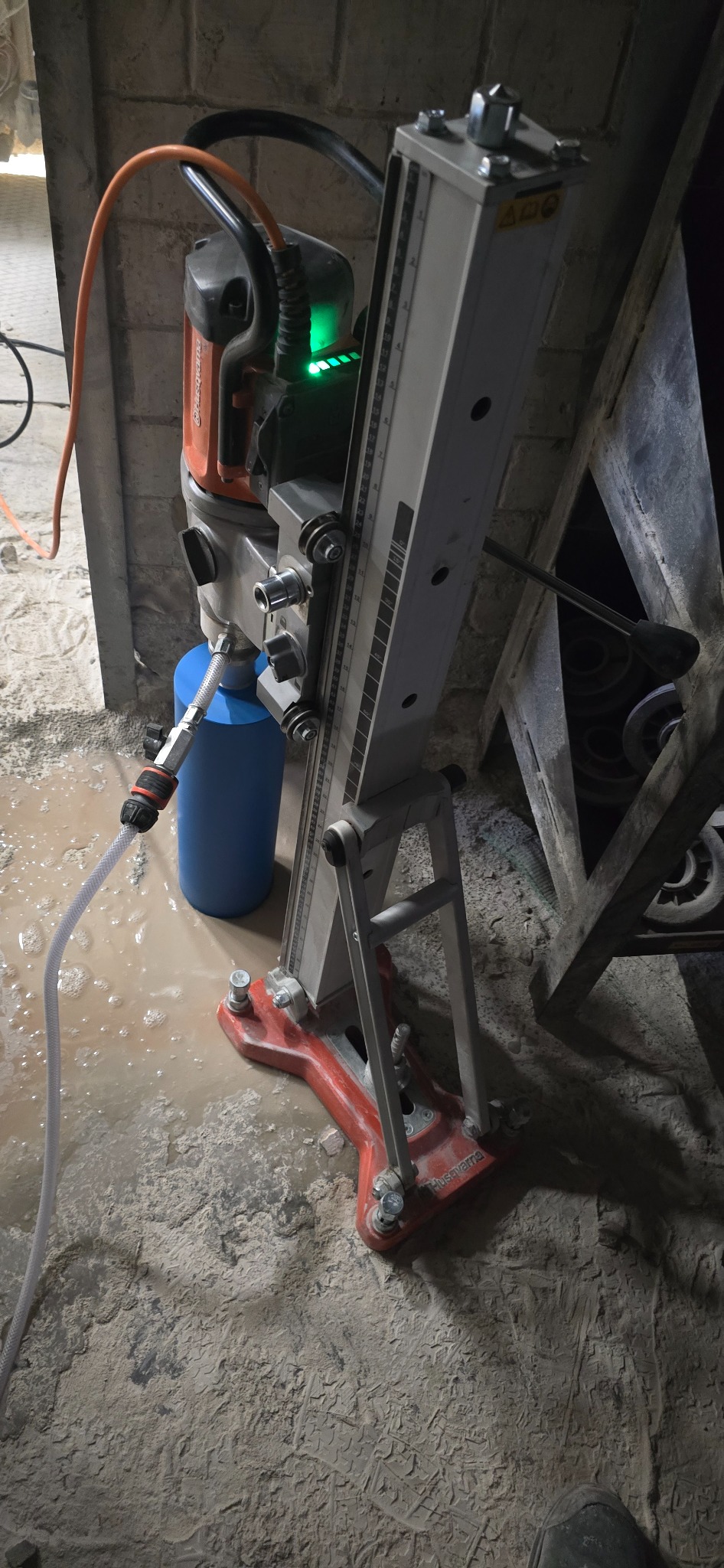 Wiertnica diamentowa Husqvarna z niebieskim zbiornikiem na wodę, ustawiona na zalanym, zakurzonym betonowym podłożu podczas wiercenia rdzeniowego.
