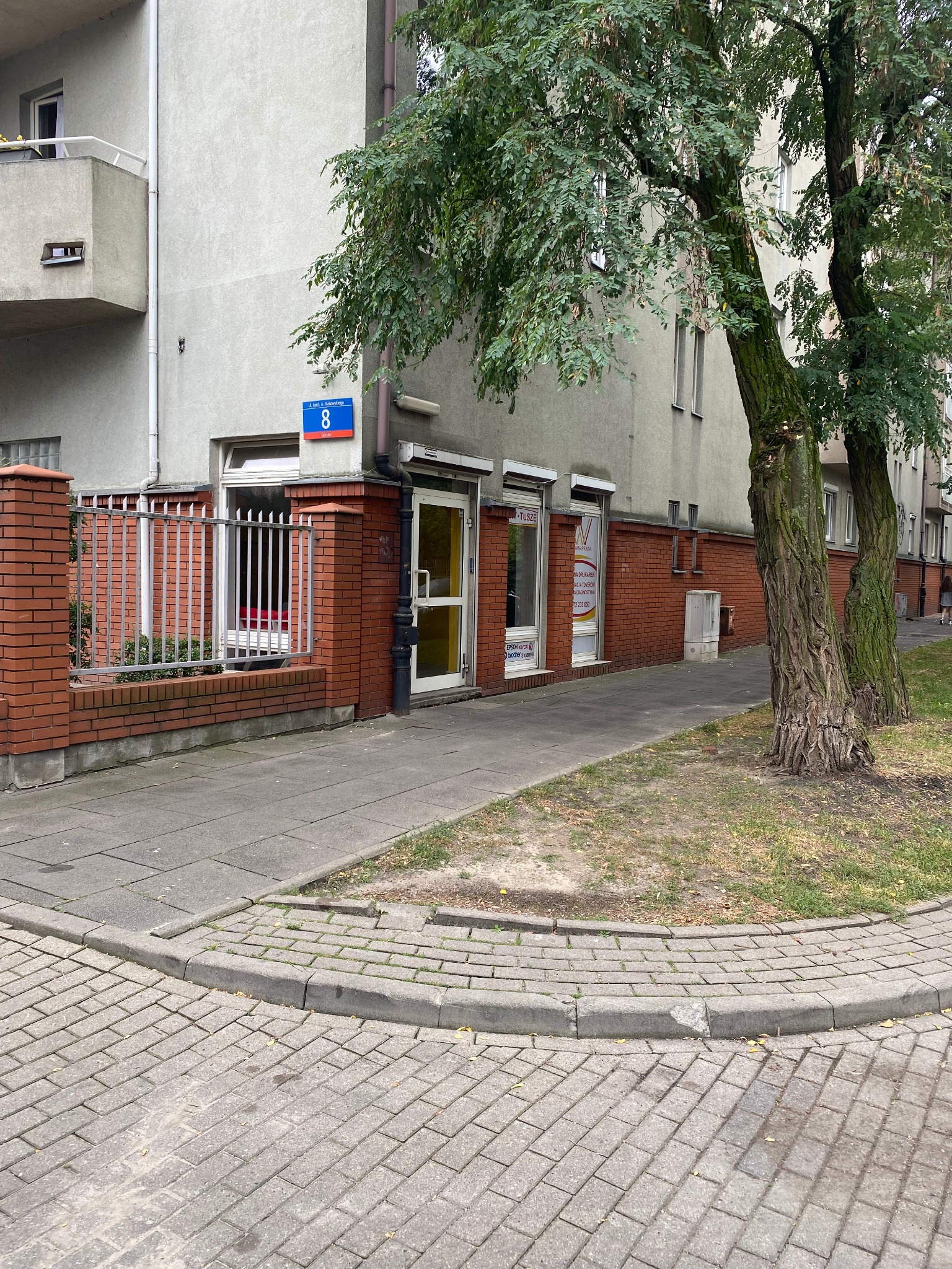 Fasada budynku z cegły z numerem 8 i szyldami reklamowymi punktu usługowego, widoczne drzewa