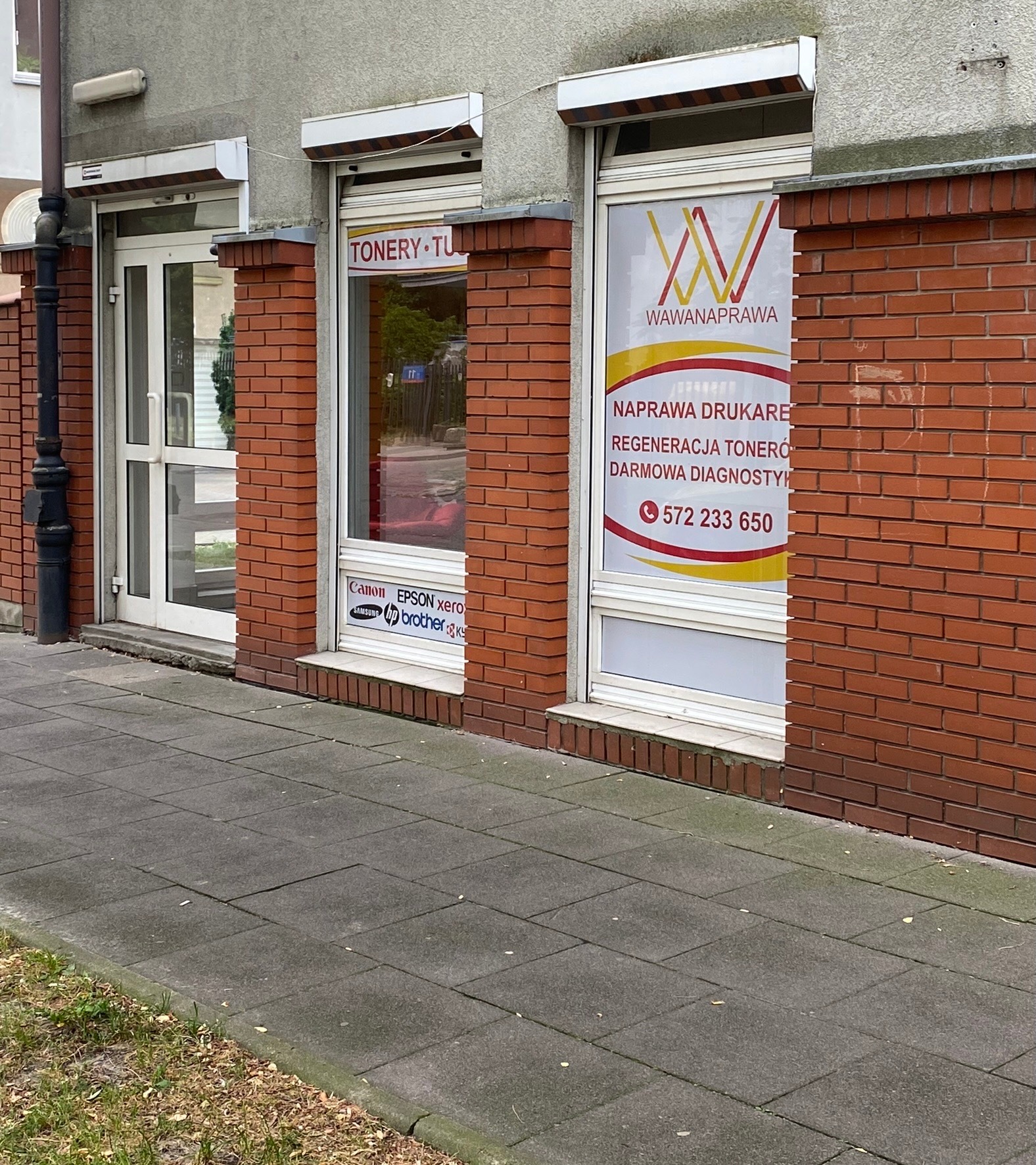 Fasada budynku w Warszawie z witrynami firmy Wawanaprawa oferującej naprawę drukarek i regenerację tonerów, widoczne logo firmy oraz naklejki z logo producentów sprzętu biurowego.
