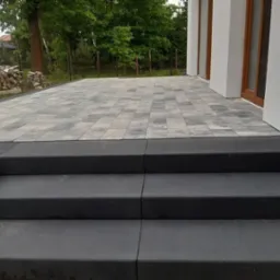 Wykończony taras z kostki brukowej w odcieniach szarości z trzema czarnymi stopniami prowadzącymi do budynku. W tle widoczne ułożone drewno opałowe i zieleń drzew.