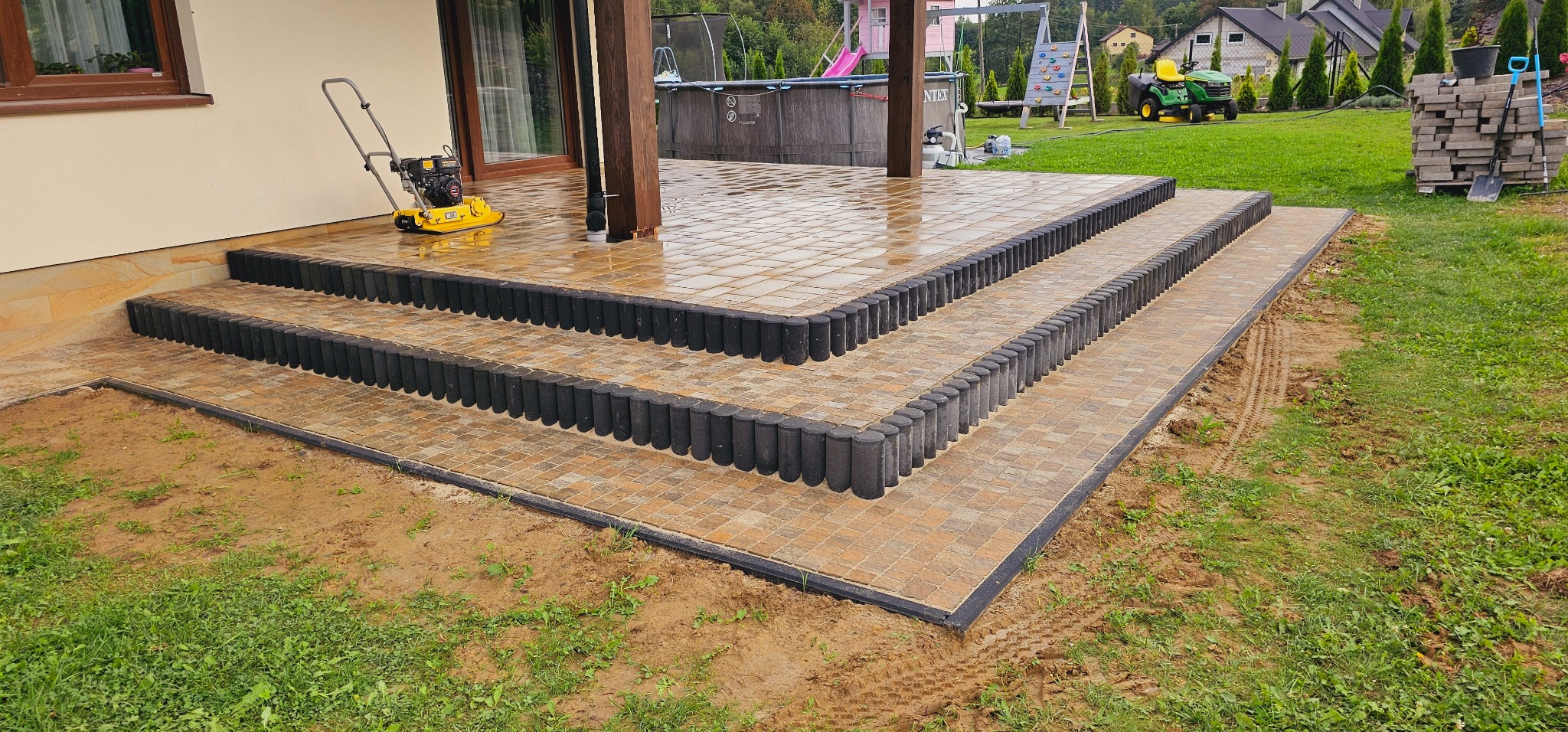 Trzy stopnie wejściowe do domu wyłożone kostką brukową w odcieniach beżu i brązu, obrzeża z ciemnych palisad, żółta zagęszczarka wibracyjna obok wejścia, w tle trawnik, basen i plac zabaw.