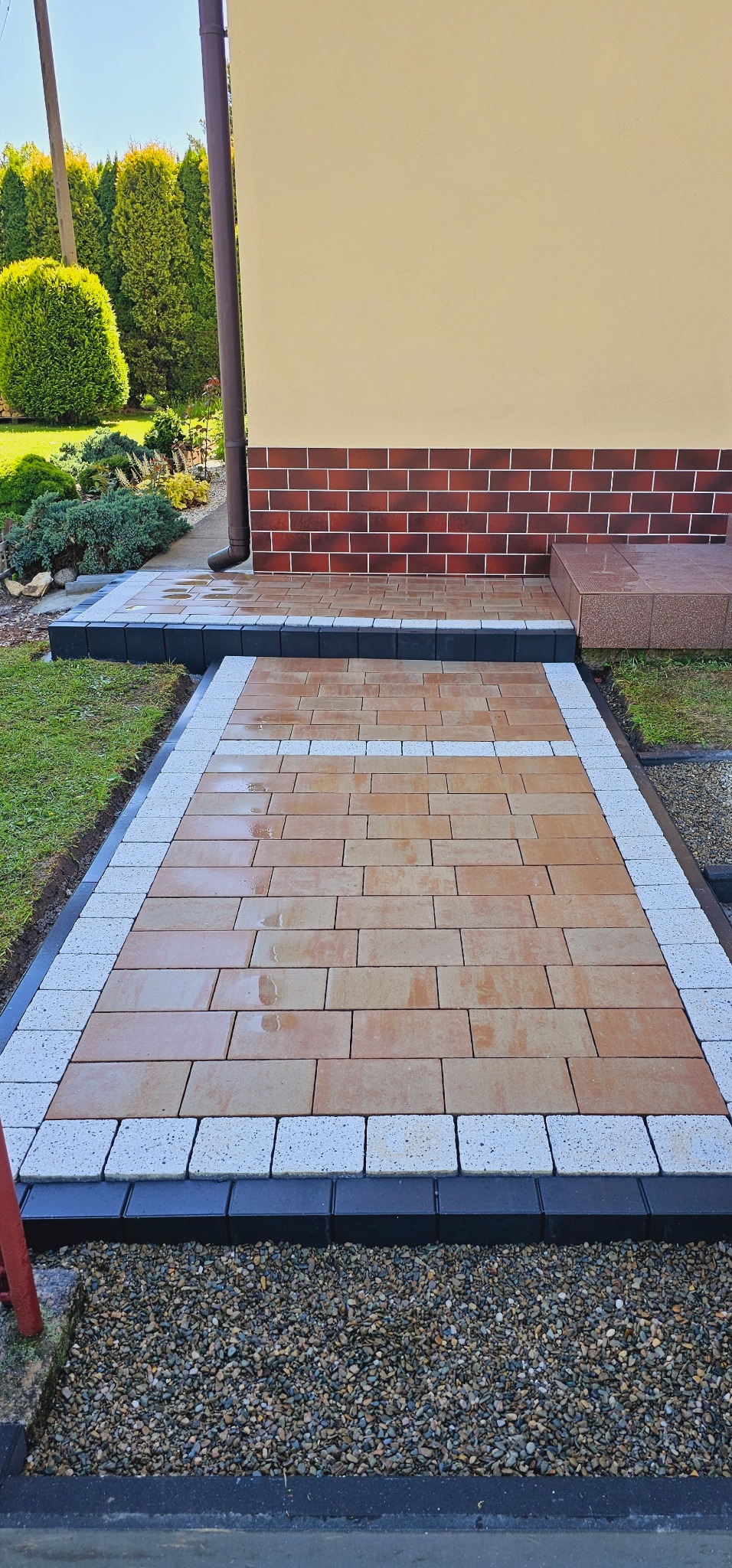 Ułożona kostka brukowa w odcieniach brązu, obramowana jasnoszarą kostką i czarnym obrzeżem, prowadząca do wejścia domu z ceglanym cokołem i beżową elewacją, z widocznymi tujami w tle.