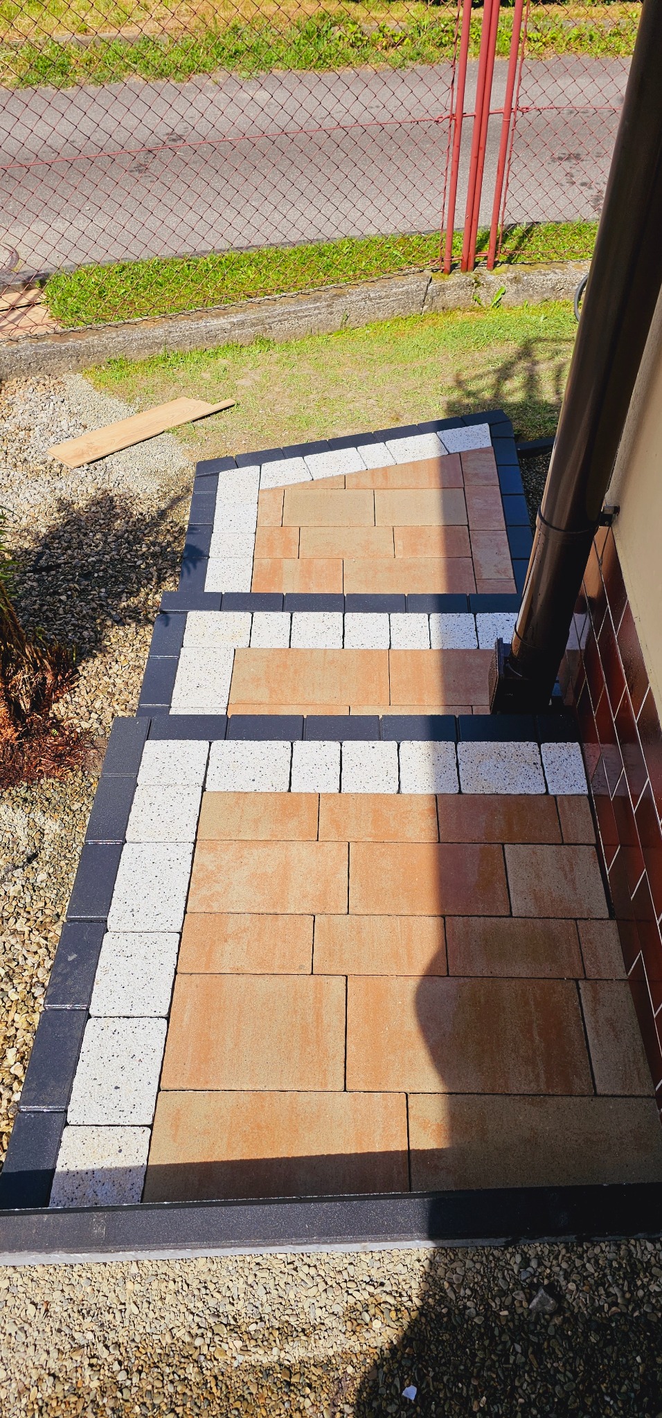 Schody wejściowe wyłożone kostką brukową w odcieniach beżu, obramowane czarną i białą kostką, widoczne z góry, z rynną i fragmentem elewacji budynku w tle.