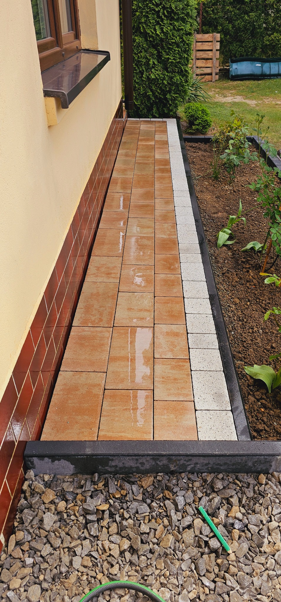 Ścieżka z brązowej kostki brukowej obok domu, obrzeże z jasnej kostki, podsypka z kamyków, widoczny zielony wąż ogrodowy i ołówek.