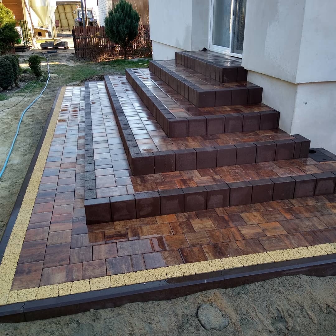 Schody wejściowe wyłożone brązową kostką brukową z żółtą obwódką, prowadzące do białego budynku z widocznym oknem.