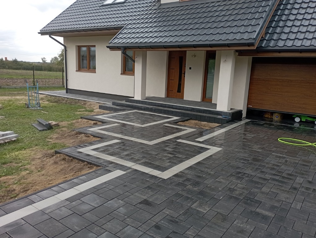 Nowoczesny podjazd z ciemnoszarej kostki z białymi akcentami, prowadzący do domu z brązową bramą garażową i schodami wejściowymi, widoczny fragment trawnika i narzędzia.