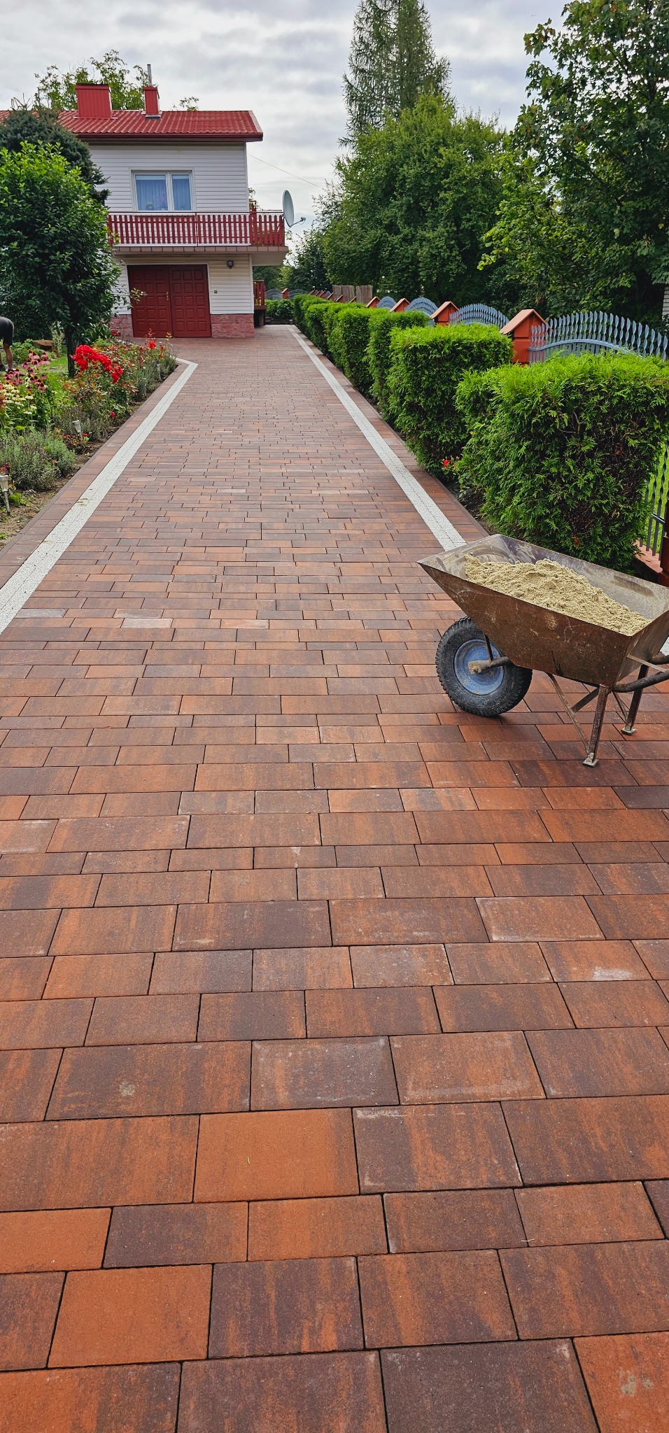 Ułożona ścieżka z brązowej kostki brukowej prowadząca do domu, z białymi obrzeżami i taczką wypełnioną piaskiem po prawej stronie.
