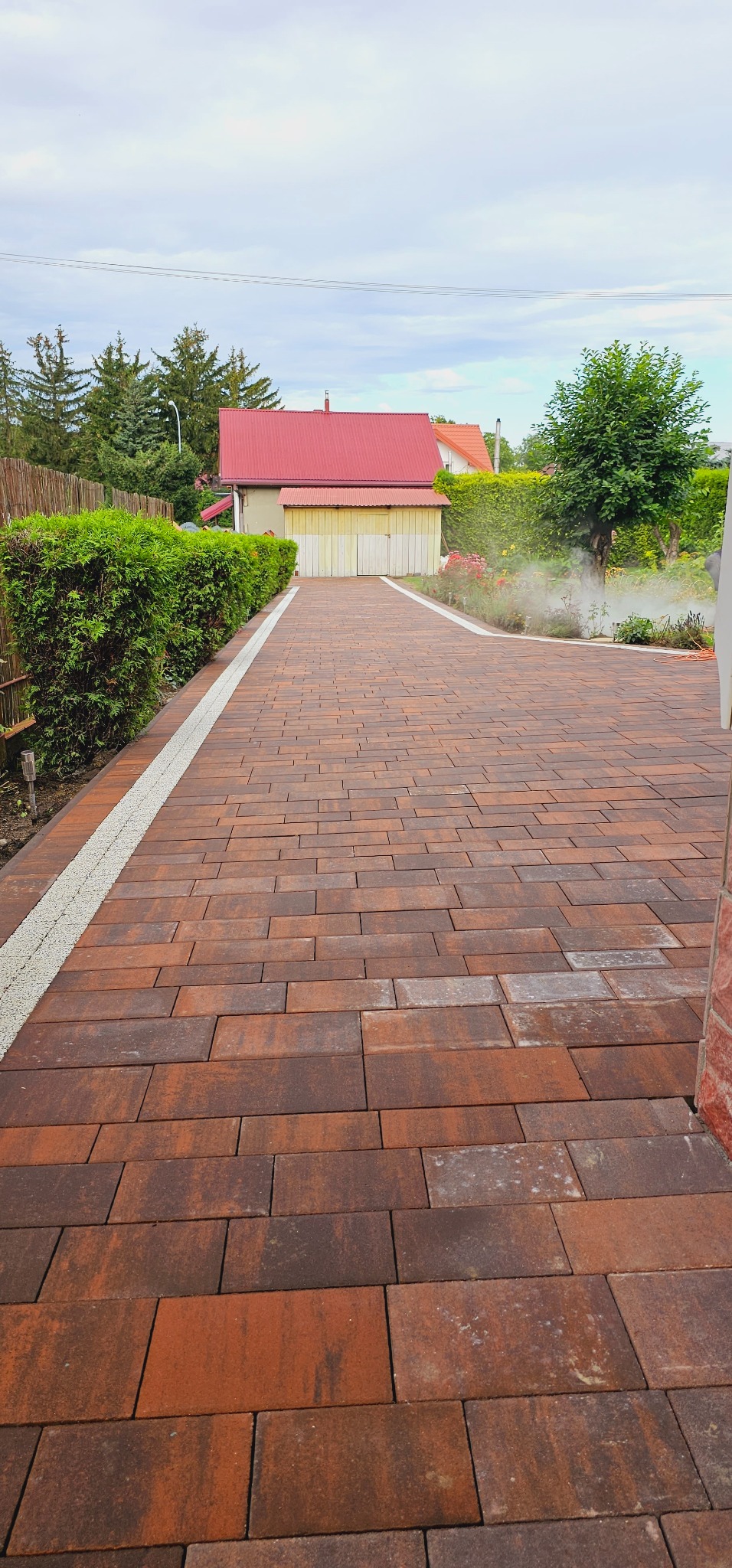 Ułożona z brązowo-czerwonej kostki brukowej ścieżka prowadząca do garażu z czerwonym dachem, obrzeża ścieżki wysypane drobnym, jasnym żwirem.