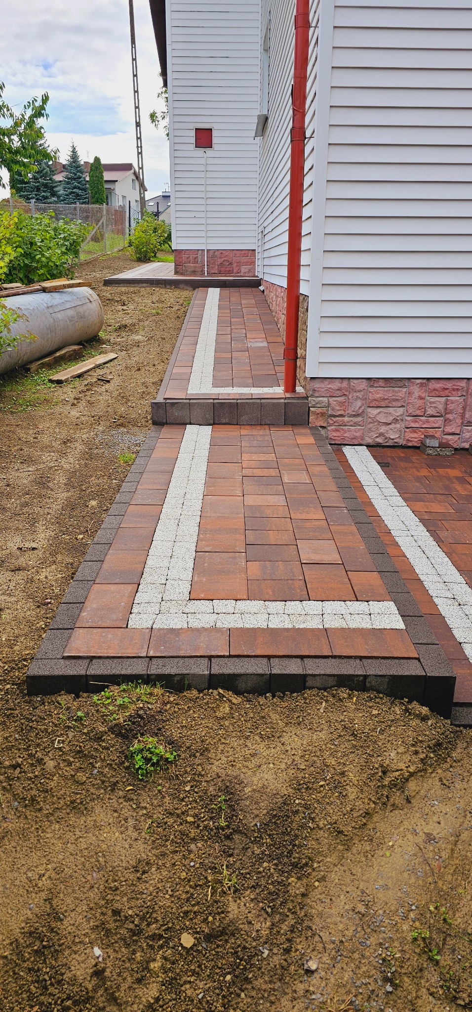 Ułożona ścieżka z brązowej kostki brukowej z jasnymi paskami dekoracyjnymi, prowadząca wzdłuż białego domu z czerwoną rynną, widok z poziomu gruntu.