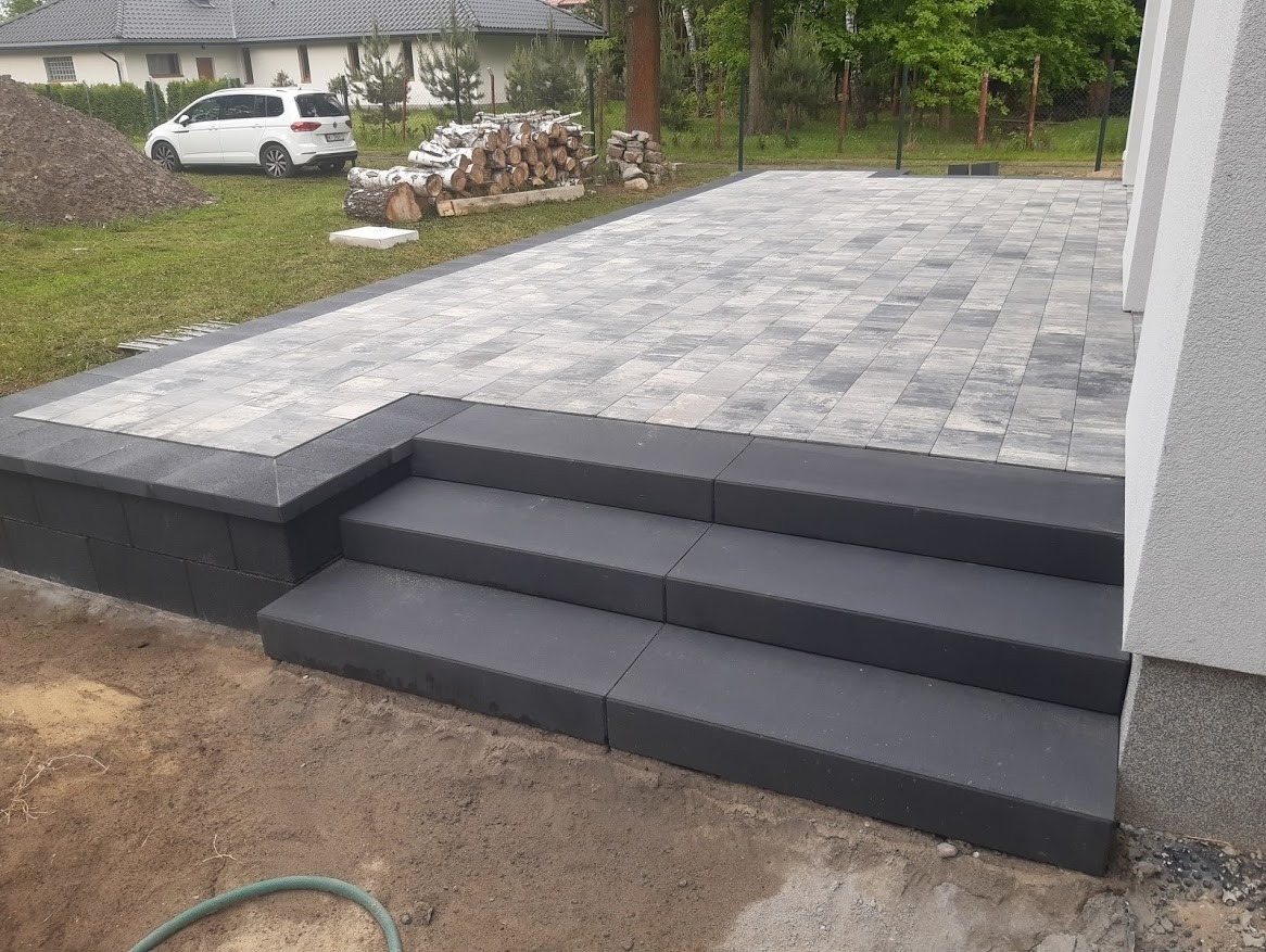 Ułożona szara kostka brukowa na tarasie z trzema stopniami, widok na trawnik, samochód i stos drewna w tle.
