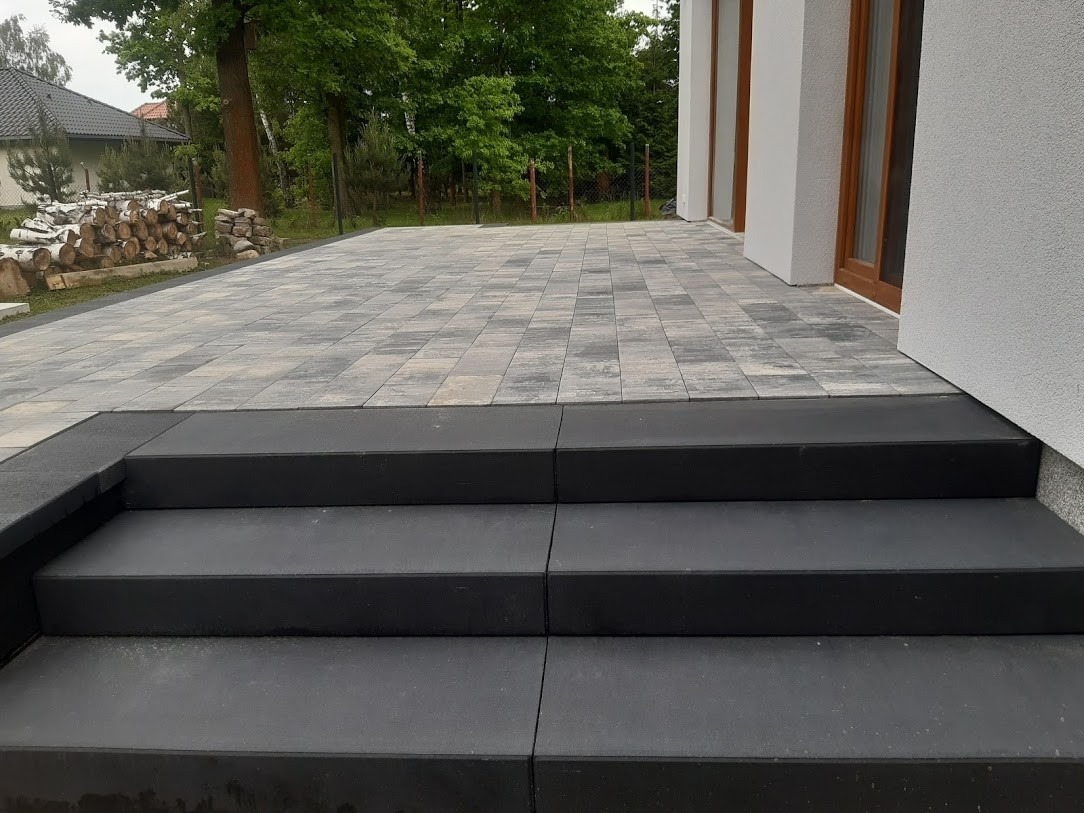 Wykończony taras z kostki brukowej w odcieniach szarości z trzema czarnymi stopniami prowadzącymi do budynku. W tle widoczne ułożone drewno opałowe i zieleń drzew.