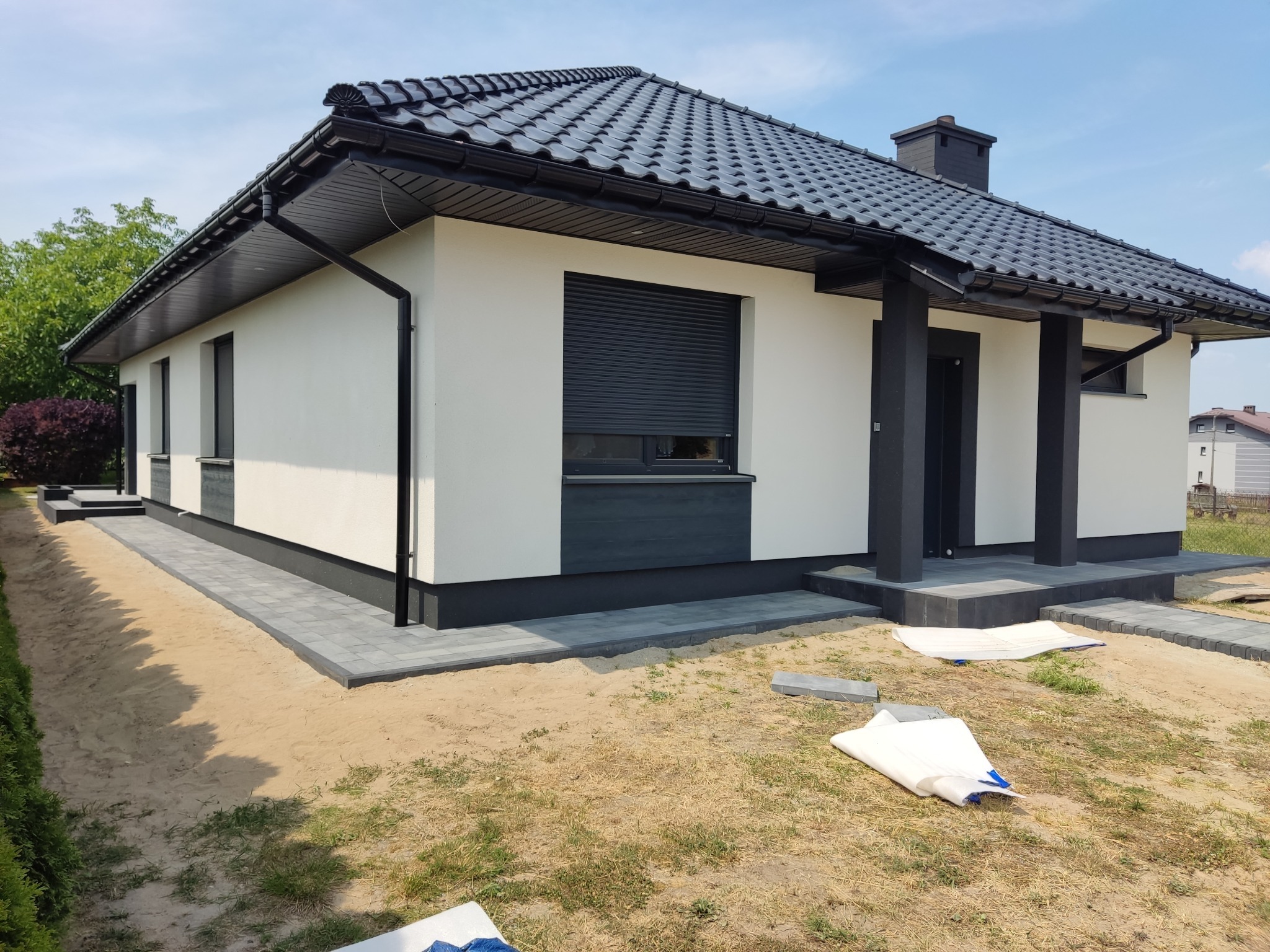 Nowoczesny dom jednorodzinny z ciemną dachówką i szarą kostką brukową wokół budynku, teren wokół domu w trakcie zagospodarowania, widoczne zabezpieczenia na ziemi.