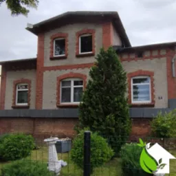 Odnowiona elewacja dwukondygnacyjnego domu z czerwonej cegły i szarego tynku w Wolsztynie, widok zza zielonego ogrodzenia i ozdobnego krzewu.