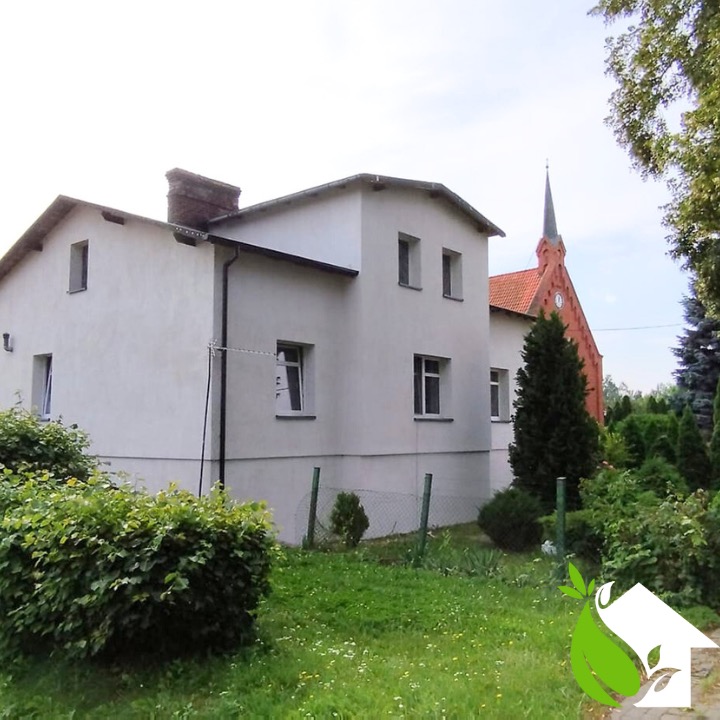 Biały budynek z ceglanym kominem i widoczną wieżą kościoła w tle, otoczony zielenią.