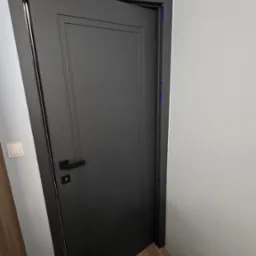 Ciemne drzwi wewnętrzne z prostokątnymi frezami, lekko uchylone, z widocznymi niebieskimi taśmami zabezpieczającymi ościeżnicę podczas montażu.