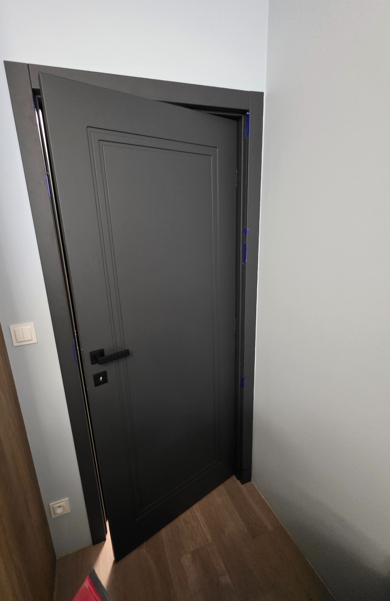 Ciemne drzwi wewnętrzne z prostokątnymi frezami, lekko uchylone, z widocznymi niebieskimi taśmami zabezpieczającymi ościeżnicę podczas montażu.