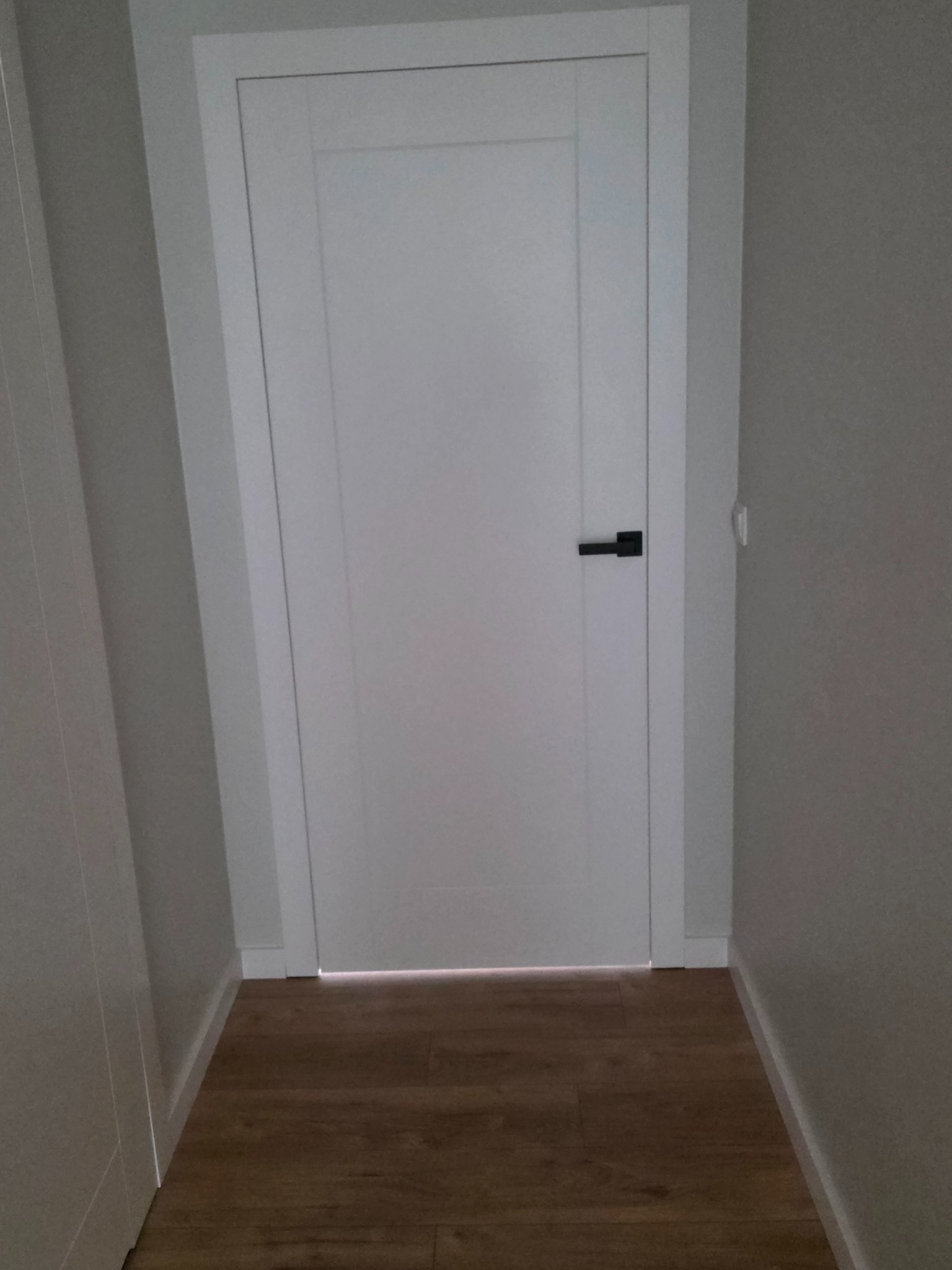 Białe drzwi wewnętrzne z czarną klamką zamontowane w szarej ścianie, widoczne panele podłogowe. Minimalistyczny design.