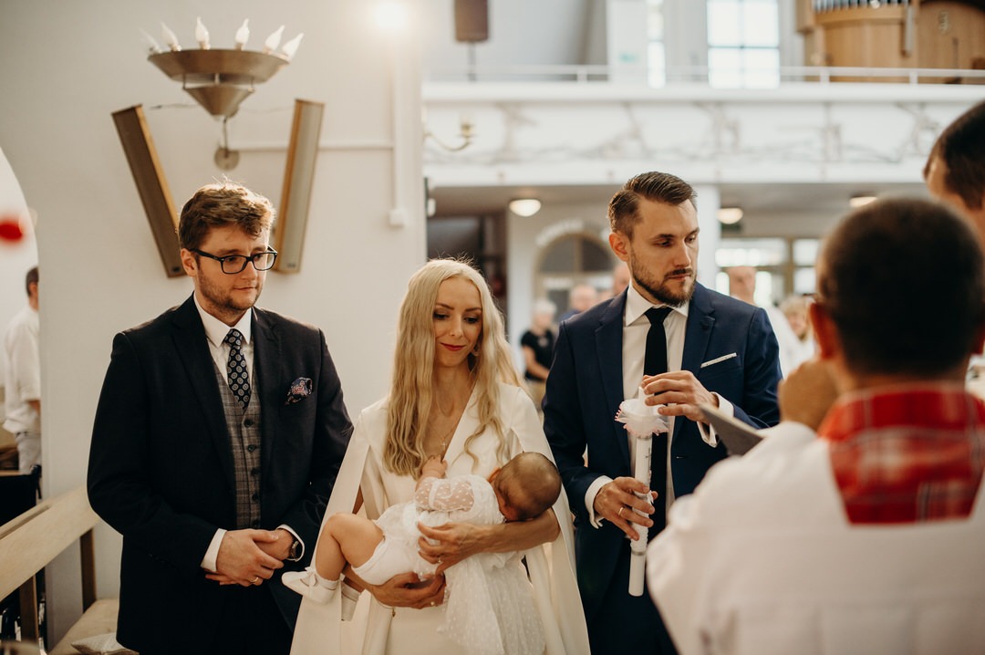 Reportaż z Chrztu Świętego: matka w białej sukni trzyma dziecko w białym ubranku, obok ojciec chrzestny z zapaloną świecą, w tle ksiądz i inni goście w kościele.