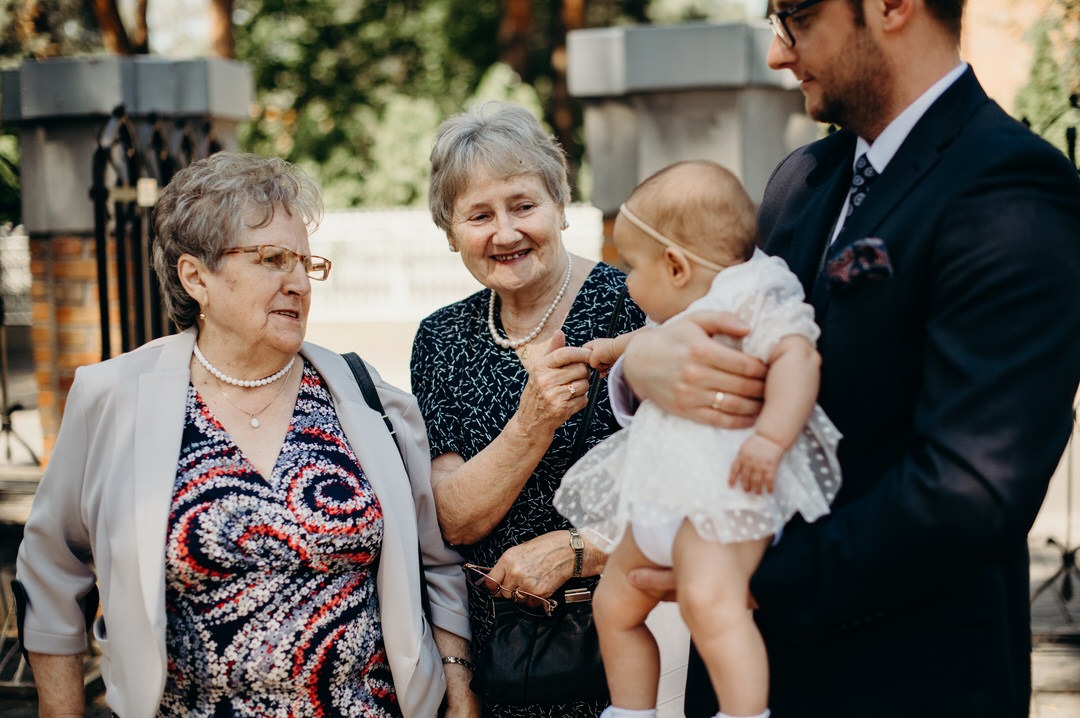 Uśmiechnięta rodzina podczas Chrztu Świętego: mężczyzna w garniturze trzyma na rękach ubraną w białą sukienkę dziewczynkę, obok stoją dwie starsze kobiety w eleganckich strojach.
