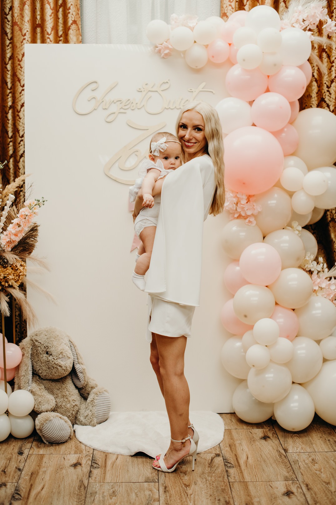Uśmiechnięta blondynka w białej sukience trzyma na rękach niemowlę z kokardą, na tle dekoracji z białych i różowych balonów oraz napisu 'Chrzest Święty', obok pluszowy królik.