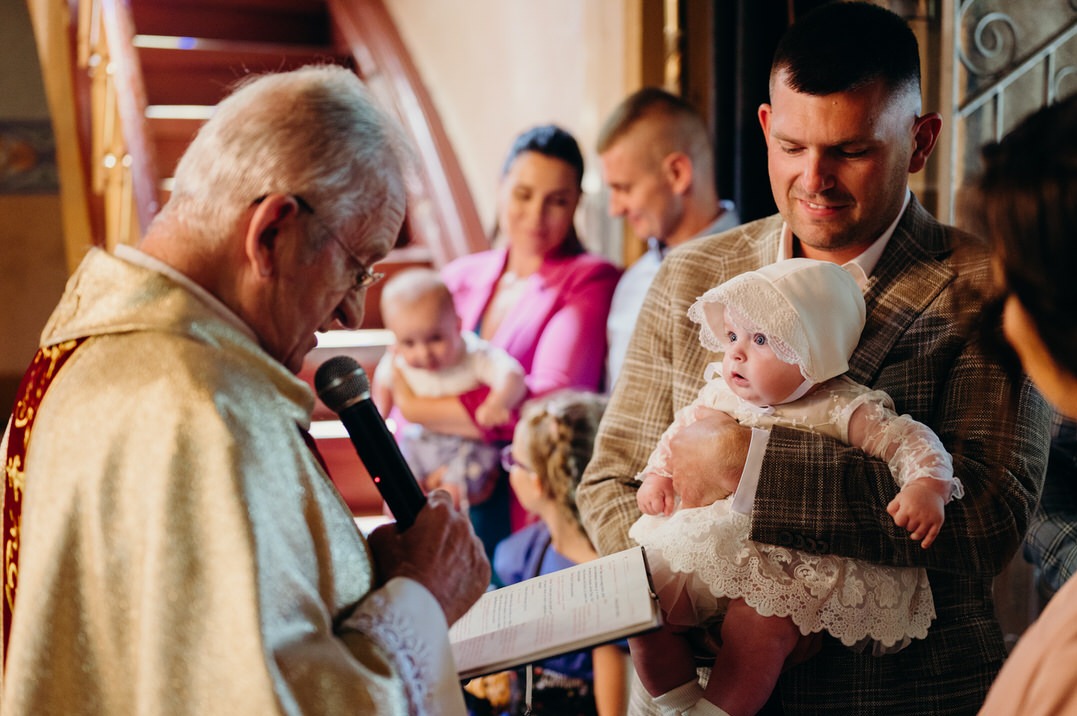 Ksiądz w bogato zdobionej szacie, trzymający mikrofon i otwartą księgę, błogosławi dziecko ubrane w białą sukienkę i czepek, trzymane przez mężczyznę w brązowej marynarce na tle rodziny podczas...