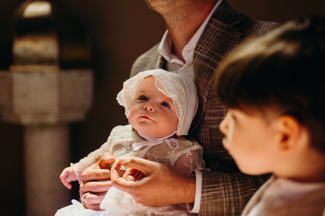 Uroczy niemowlak w białej koronkowej sukience i czapeczce, trzymany na rękach mężczyzny w kraciastej marynarce, z fragmentem drugiego dziecka w tle, w kościelnym wnętrzu.