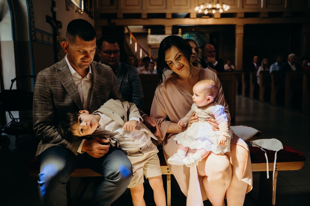 Rodzina siedzi w ławce kościoła podczas Chrztu Świętego: tata w kraciastej marynarce trzyma syna w beżowym garniturze, mama w pudrowej sukience trzyma niemowlę w białej sukience z koronki.