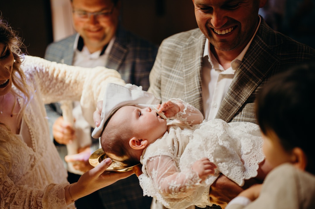 Uśmiechnięty ojciec trzyma w ramionach niemowlę w białej, koronkowej sukience podczas chrztu, gdy kapłan polewa główkę wodą ze złotej miseczki, a w tle widać uśmiechniętych gości.