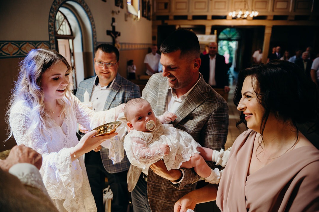 Uśmiechnięty ojciec trzyma niemowlę w białej sukience z koronki podczas Chrztu Świętego, obok matka chrzestna z naczyniem na wodę święconą i inni goście w tle w kościele.