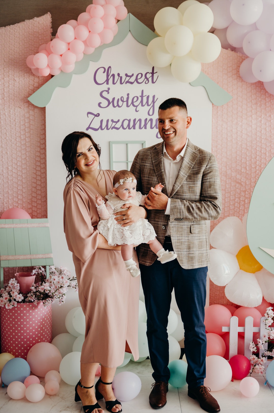 Uśmiechnięta rodzina pozuje na tle dekoracji z okazji Chrztu Świętego Zuzanny, z balonami i napisem na białej ściance w kształcie domku.