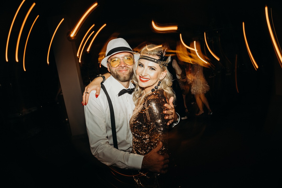 Para w strojach z lat 20. na tle rozmazanych świateł podczas przyjęcia urodzinowego; mężczyzna w białym kapeluszu i żółtych okularach obejmuje kobietę w złotej sukience cekinowej i opasce na głowie...