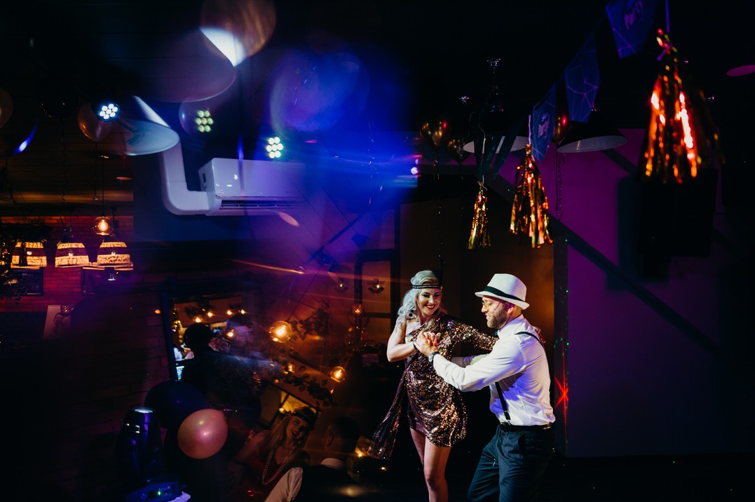 Para tańczy w stylu lat 20. na tle dekoracji urodzinowych, oświetlona reflektorami i wiszącymi lampami.